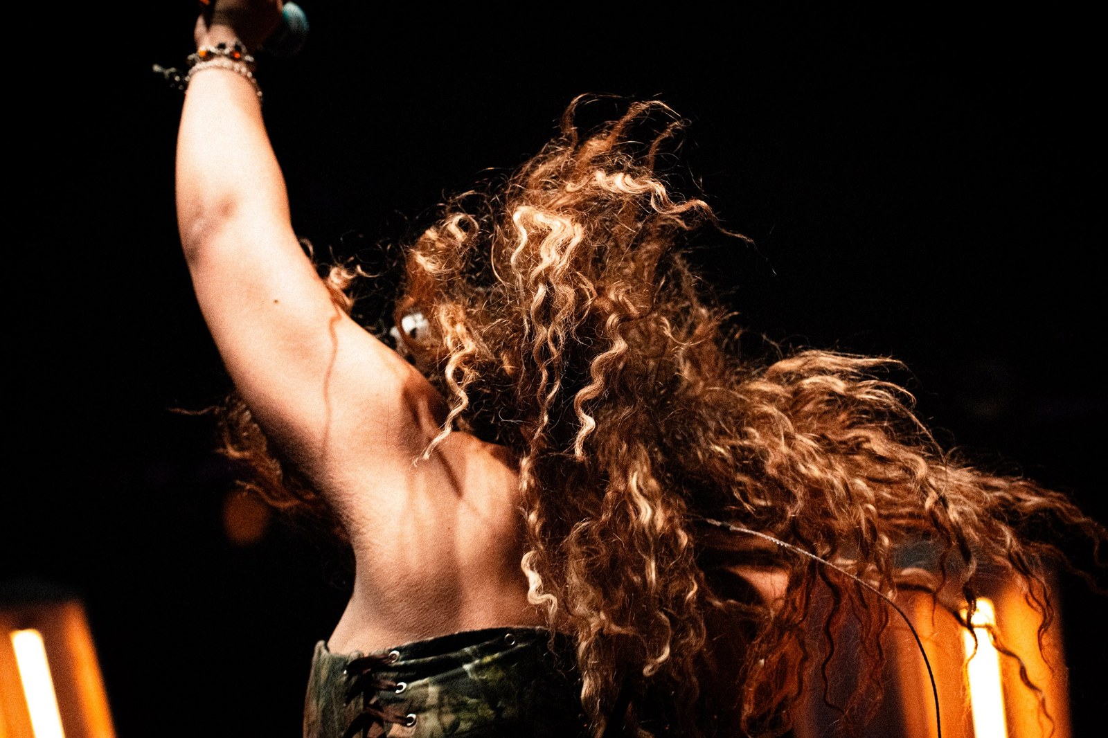 Singer seen from behind, arm raised, curly hair flying in orange light