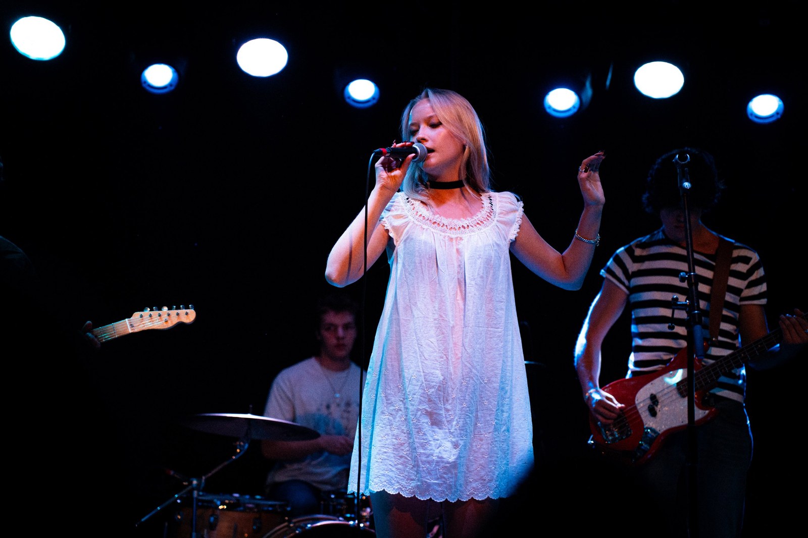 Singer in a white dress at the mic, band behind her under blue stage lights