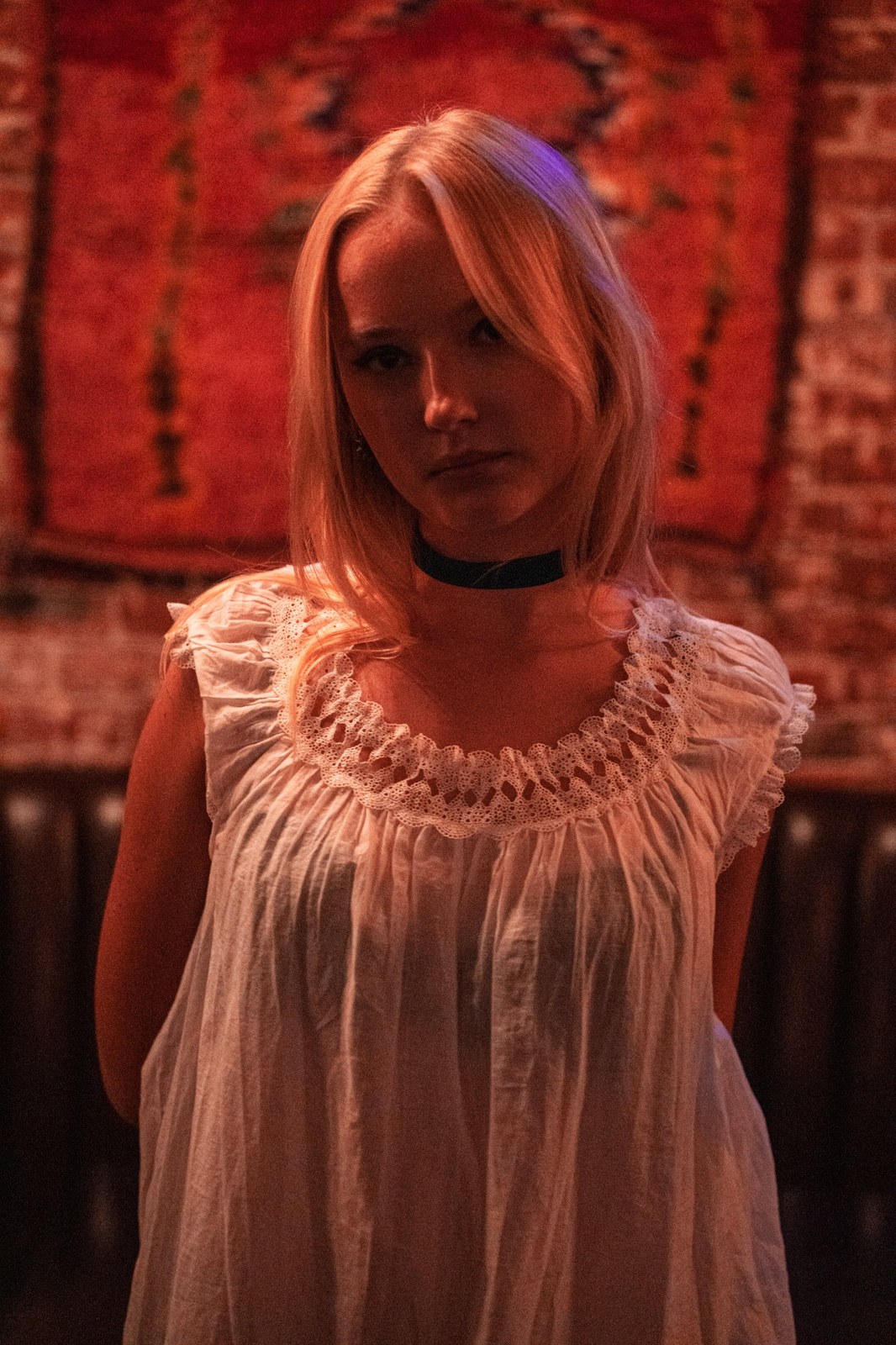 Woman in a white lace dress against a red rug and brick wall, looking at the camera