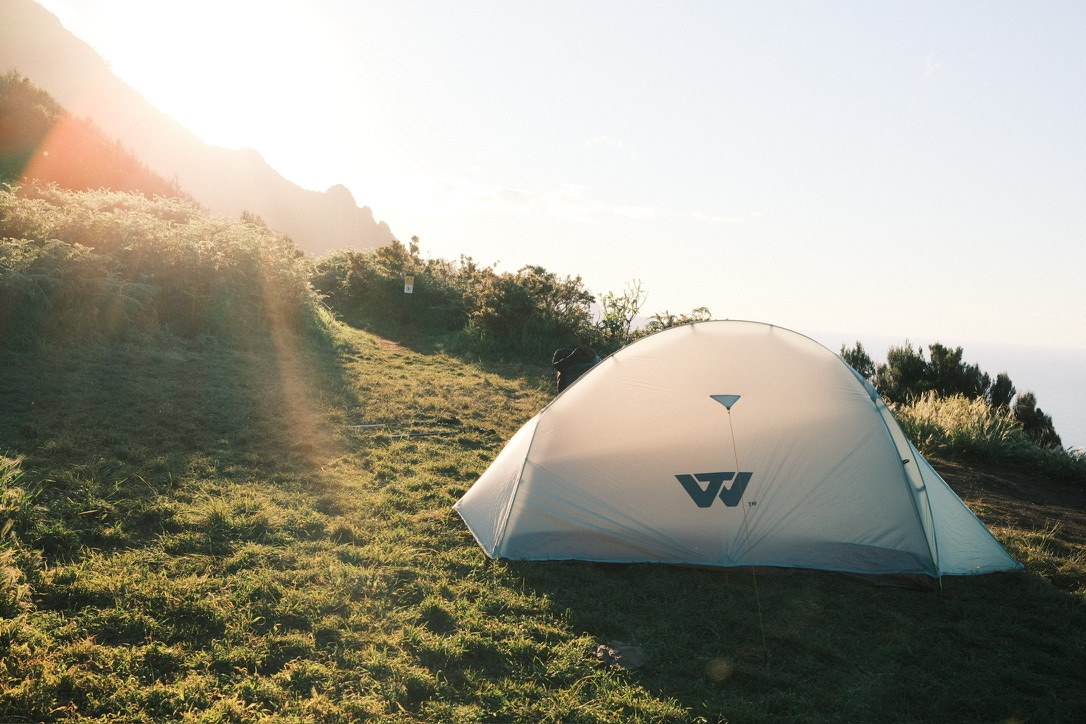 Tent pitched on a grassy ridge at sunrise, sun flaring over the cliffs